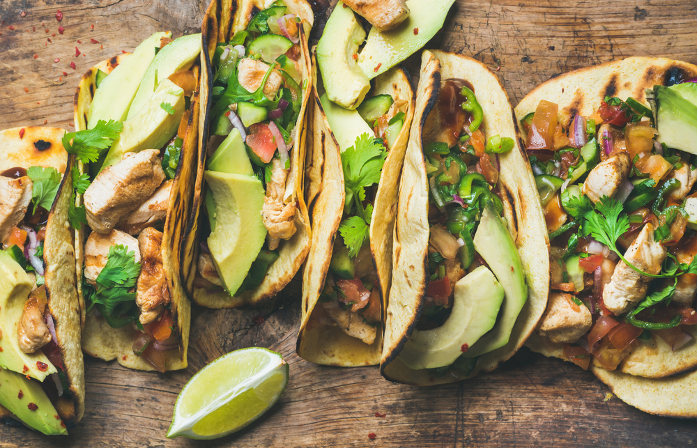 Tacos with grilled chicken, avocado, fresh salsa sauce and limes over rustic wooden background, top view.