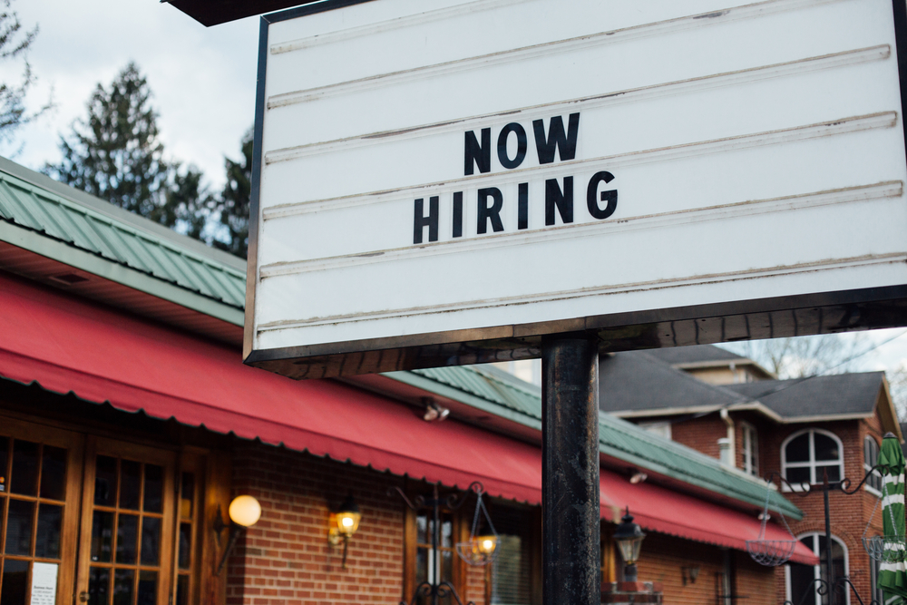 Typical american now hiring sign in black letters over the white background with local small business building